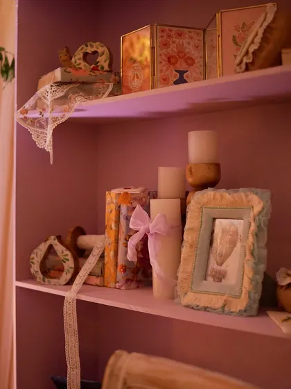 A shelf painted in a soft lavender hue, styled with candles, books, and framed pictures.