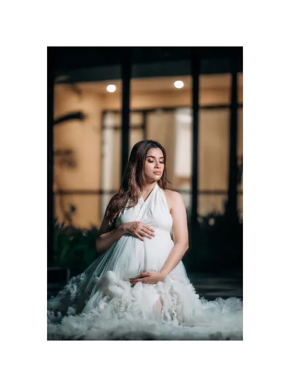 A peaceful and serene portrait of a mother-to-be seated outdoors at night, cradling her bump. The soft lighting creates a beautiful glow, highlighting her white ruffled dress.
