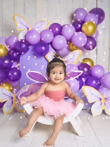 A sweet toddler dressed as a butterfly, smiling brightly for her milestone photoshoot. The purple and gold balloon arch and butterfly cutouts create a magical, whimsical setting.