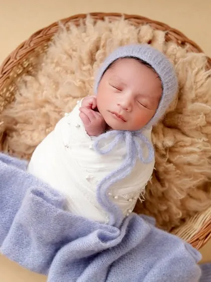 This little one is wrapped snugly in a white swaddle and a blue knit blanket, looking cozy and content in a fur-lined basket.