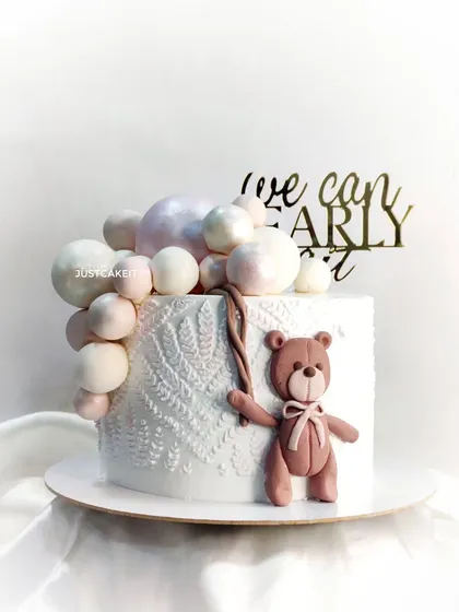 A "We Can Bearly Wait" baby shower cake. The design is modern and sweet, with a knitted texture on the cake and a cute teddy bear holding a balloon cascade.
