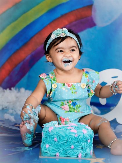That "I can't believe this is all for me" smile! This little girl is loving every moment of her rainbow-themed cake smash, with blue frosting and a whole lot of happiness.