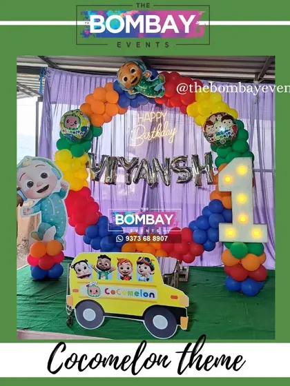 This Cocomelon theme uses the full rainbow of colors in its circular balloon arch. The setup is completed with a light-up number '1', a Cocomelon bus, and a JJ cutout.