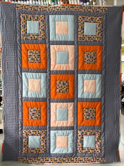 A modern take on a block quilt with a grey, orange, and blue color palette. The border features a geometric maze-like quilting pattern.