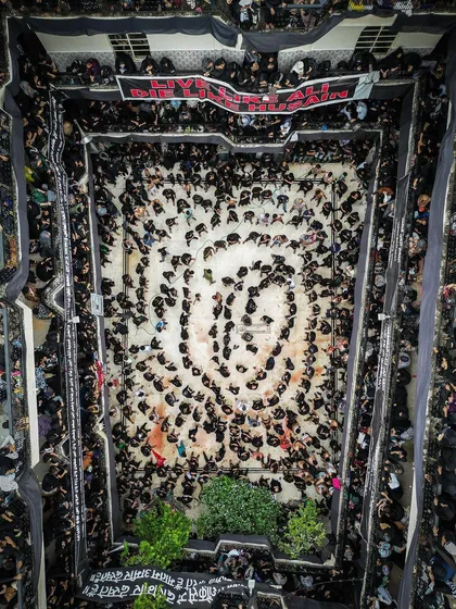 A wide, top-down aerial shot of a Muharram procession, showing participants forming a massive, swirling circle of devotion. This perspective reveals the scale and organized pattern of the ritual, which is impossible to see from the ground.