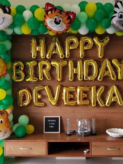 A jungle-themed decoration for a first birthday at home. The setup features a balloon frame with animal foils around a TV console, with the name 'DEVEEKA' in large foil letters.