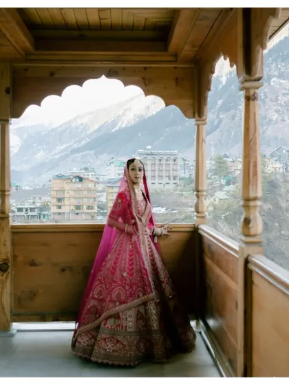The bride, framed by a traditional wooden balcony, looks out towards the Manali valley. Her vibrant pink lehenga is a stunning splash of color against the earthy tones of the mountains and architecture.