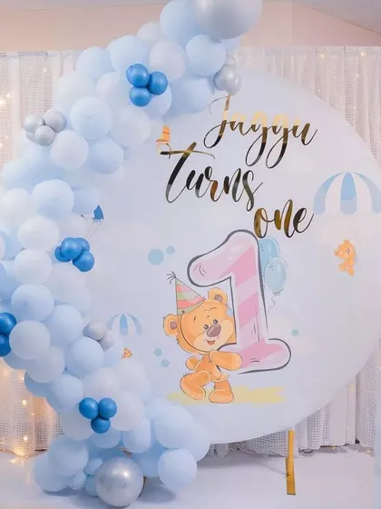 A charming teddy bear first birthday backdrop. This simple and sweet setup features a large circular print with a cute bear illustration, framed by a blue and white balloon arch.