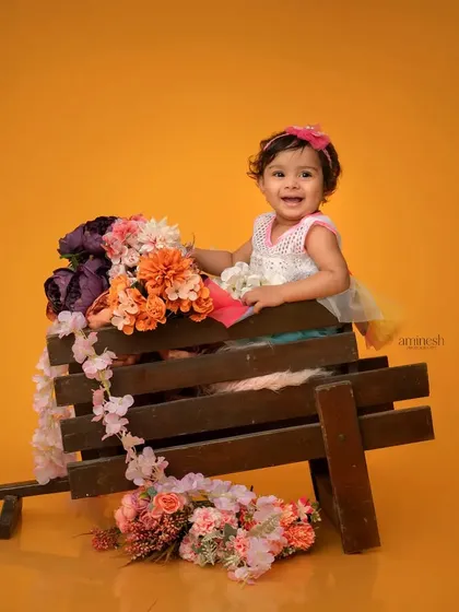 A happy baby girl sits in a wooden wheelbarrow decorated with flowers. The bright yellow background and her big smile make this an incredibly cheerful photo.