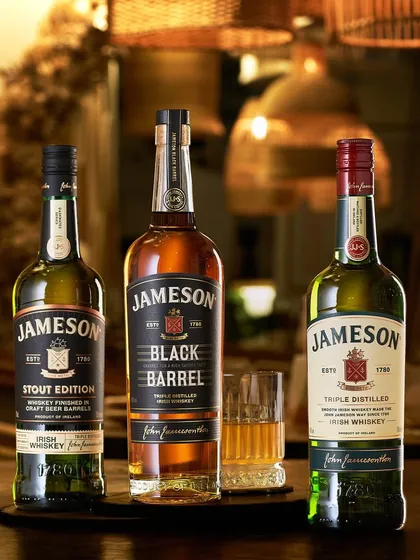 The Jameson family. This shot showcases three different Jameson expressions in a warm, inviting bar environment. The background is softly blurred to keep the focus on the distinct character of each bottle.
