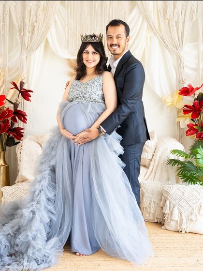 A full-length portrait of a glamorous couple. The mom-to-be wears a stunning blue gown while her partner stands proudly by her side.