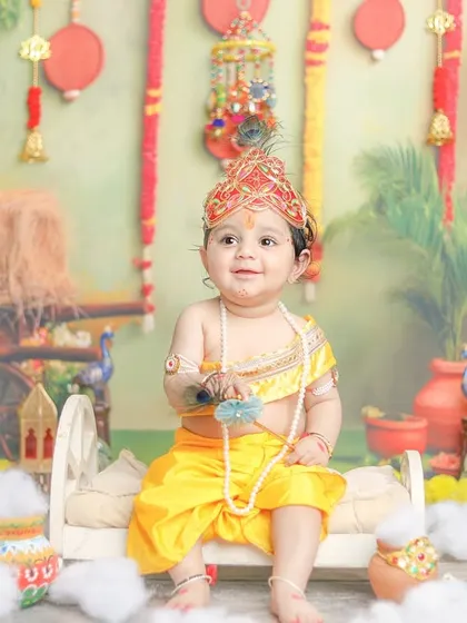 This little Krishna is ready for his close-up, sitting on a miniature bed amidst a festive setup.