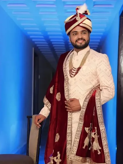 A full-length shot of the groom in his elegant ivory sherwani and red velvet shawl. The complete styling creates a regal and memorable wedding day appearance.