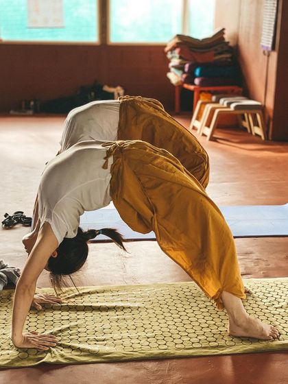 A student practices a variation of a forward bend, demonstrating focus and flexibility. The warm light of the shala creates a peaceful atmosphere for deep, introspective practice.