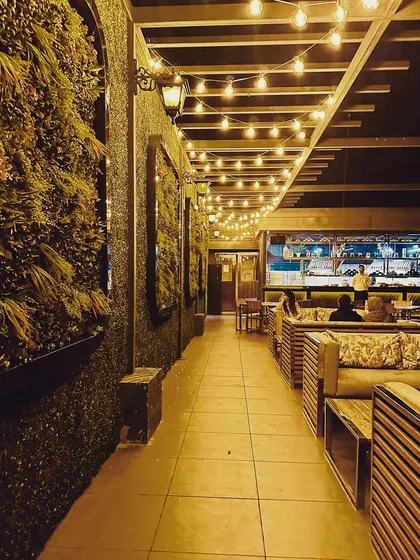 An inviting seating area at a rooftop cafe project. I used a combination of natural-looking green walls, warm string lights under a pergola, and comfortable wooden furniture to create a cozy and rustic atmosphere for guests to enjoy.