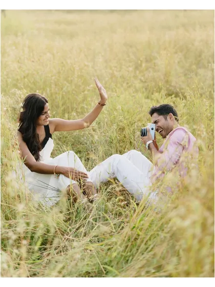 A wider angle of their playful polaroid shoot. It shows the full scene, capturing their interaction within the beautiful, natural setting of the tall grass field.