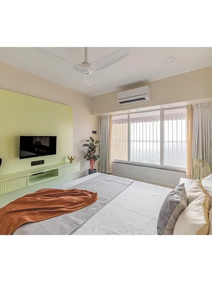 A bright and airy master bedroom in a Mumbai residence. The design uses a soft, minimal palette with a pop of pastel green in the headboard and TV unit to create a refreshing and modern feel.
