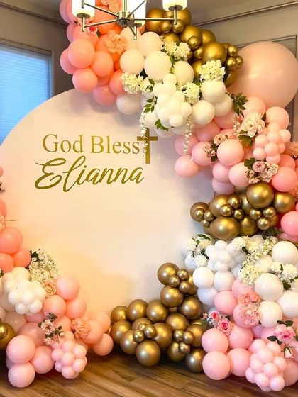 A beautiful First Holy Communion decoration with pink, gold, and white balloons. The arrangement is accented with small bouquets of fresh flowers.