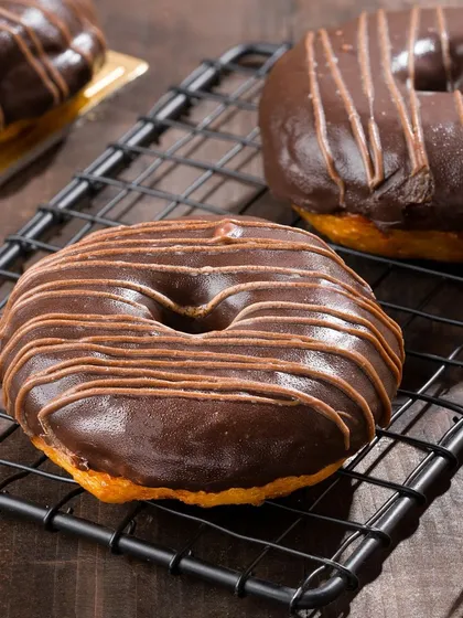 A close-up of a chocolate-drizzled donut on a cooling rack. We focus on textures and glossy finishes to make baked goods look irresistible.
