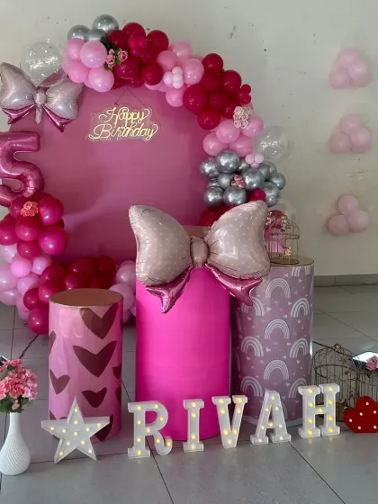 A vibrant pink birthday setup featuring a circular backdrop with a 'Happy Birthday' neon sign. The decor is personalized with marquee letters for the child's name and includes themed plinths and bow-shaped foil balloons.