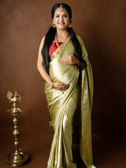 A full-length portrait of a mom-to-be in a traditional saree, standing next to a brass lamp (diya), symbolizing light and prosperity.