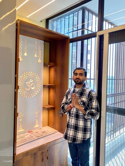 Standing with a completed mandir unit. This wooden design features a backlit acrylic panel with religious motifs and storage cabinets below.