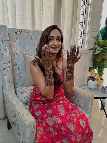 A happy bride seated in a floral chair, showing her beautiful peacock-themed mehndi.