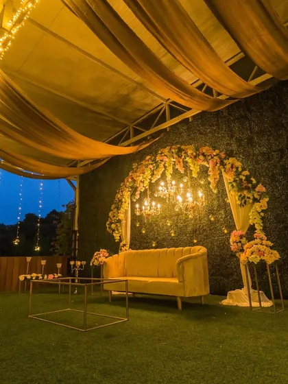 A beautiful evening stage setup with a floral arch and chandeliers against a green wall, ready for a reception or engagement.