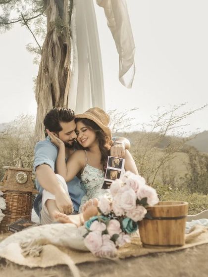 A sweet, romantic picnic scene. The couple embraces, sharing a happy moment while looking at their baby's sonogram, surrounded by flowers and a beautiful, rustic landscape.