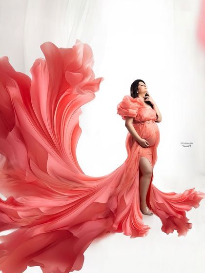 A vibrant and dynamic portrait featuring a coral-pink gown with dramatic ruffled sleeves. The fabric is tossed to create a beautiful, wing-like effect against a clean white background.