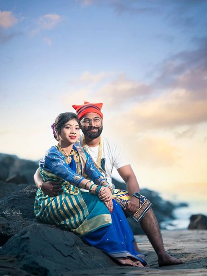 A serene portrait of the couple seated on the rocks at dusk. The bride's striped blue saree and the groom's red Koli cap stand out against the soft colors of the sky, making for a beautiful and peaceful image.