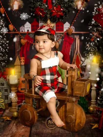 A toddler boy in a festive plaid outfit sits on a wooden tricycle in a cozy, rustic Christmas setting.