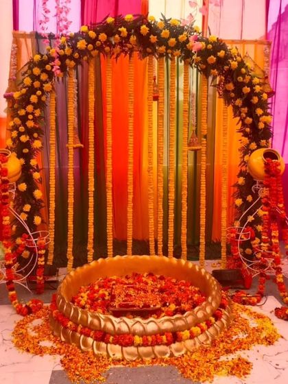 A traditional Haldi ceremony setup with a floral arch and a large brass urli. The decor uses marigold garlands and decorative matkas on stands, perfect for a home-based function.