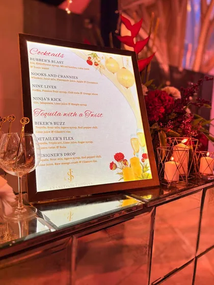 A beautifully designed, backlit cocktail menu for a wedding celebration. The menu features custom drink names like "Biker's Buzz" and "Designer's Drop," reflecting the personalities of the couple.