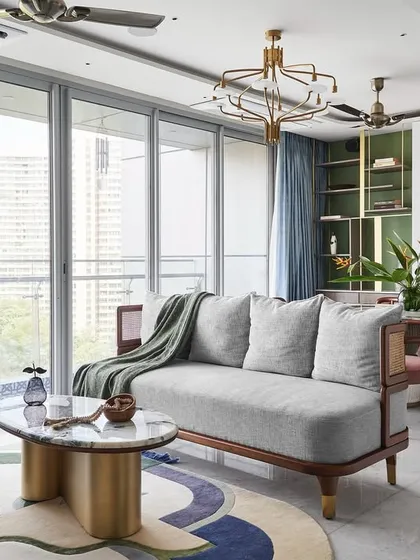 The living room is filled with natural light from the expansive windows. The custom 'Ananta' center table with its infinity-shaped brass base and the colorful rug from The Hatsu anchor the seating area.