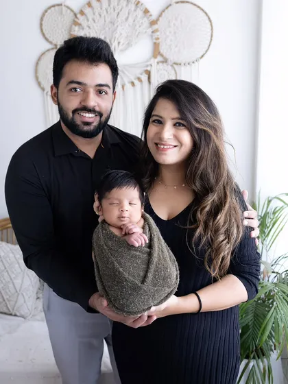 A classic family portrait with a newborn. The baby is swaddled and held securely by both parents, who are smiling warmly at the camera.