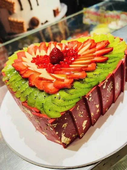 A vibrant heart-shaped fresh fruit cake, beautifully arranged with sliced kiwi, strawberries, apples, and pomegranate seeds.