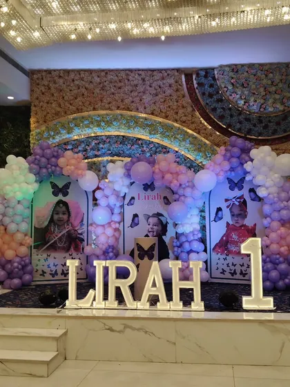 A grand stage for a first birthday party with a butterfly theme, featuring multiple photo backdrops and large light-up letters spelling the name "LIRAH".