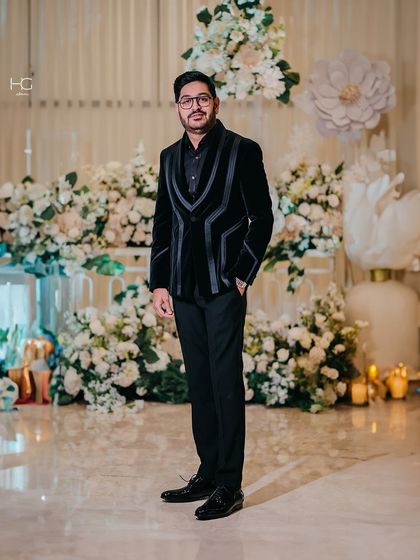 A full-length view of the client in the velvet tuxedo with geometric lapels. Set against a floral backdrop, the power of the all-black look is undeniable.