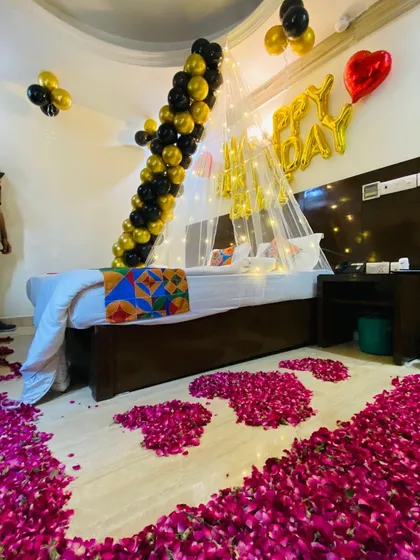 A wide shot showing the complete black and gold birthday surprise, from the balloon garland to the extensive rose petal floor decoration.