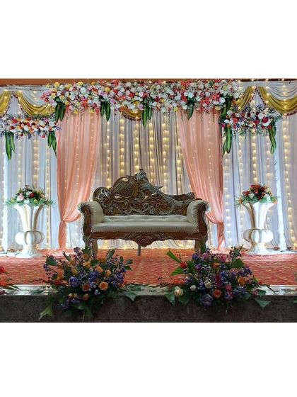 This is our classic stage decoration for a formal event like a reception or engagement. The setup includes a beautiful floral valance, elegant pink and white drapes, a regal lounge sofa, and matching flower arrangements on urns.