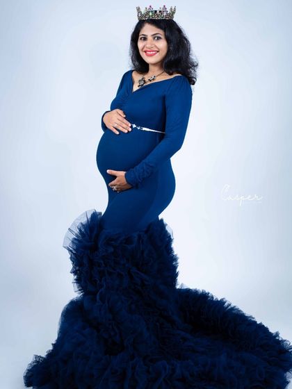 A full-length portrait in a navy blue mermaid gown with a dramatic ruffled train. The simple white background makes the color and style of the dress pop.