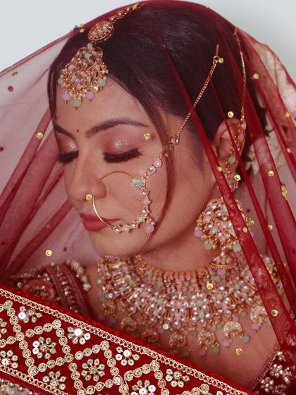 A soft and romantic bridal portrait. The focus is on the delicate eye makeup and pastel-toned jewellery, seen through a sheer red dupatta.