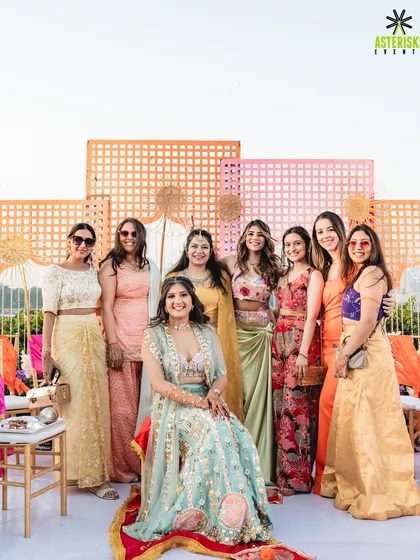 The bride poses with her squad, all dressed in vibrant outfits, against the colorful backdrop of the Mehendi decor.