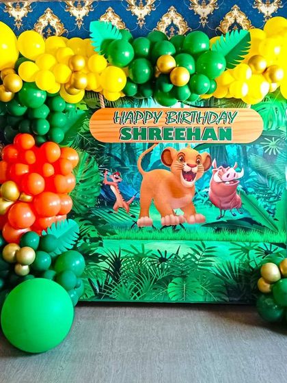 A simple jungle themed birthday decoration with a Lion King backdrop. The setup uses green, orange, and yellow balloons to create a vibrant jungle feel.