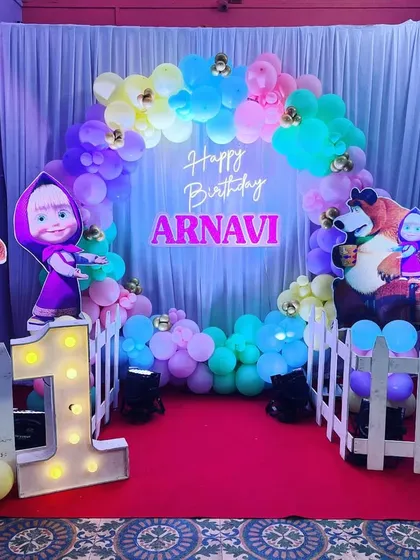A cute "Masha and the Bear" themed first birthday party. The setup includes character cutouts, a pastel balloon arch, and a light-up marquee number '1'.