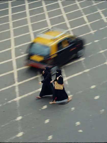 An aerial view of two women in burqas crossing the street, with a taxi blurring past. The panning motion and unique perspective create a dynamic and artistic street photograph.
