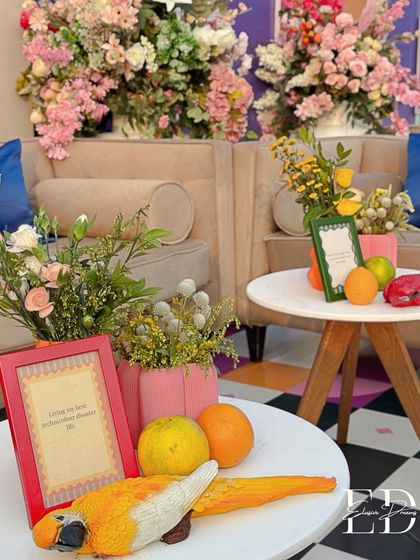 A closer look at the carnival decor, with pink vases, fresh citrus fruits, and a playful parrot statue. The background features lush pink floral arrangements, adding a touch of softness to the bold theme.