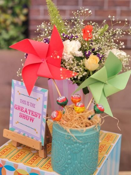 A close-up of a carnival centerpiece featuring colorful pinwheels and lollipops in a decorative pot. The "Greatest Show" sign and ticket roll base complete the theme perfectly.