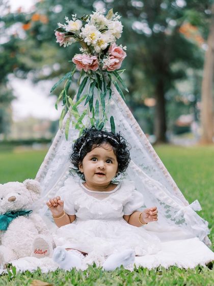 A baby girl sitting in front of a teepee, looking adorable. Outdoor milestone sessions are a wonderful way to celebrate.